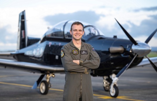 A man wearing a flight suit smiles with crossed arms in front of a small black aircraft with a propellor on the front.