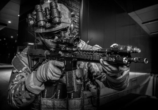 The image is black and white. The image is of a NZSAS soldier holding a weapon, pointed to the right of the image. The solider is in full uniform and wearing glasses, gloves, NVG. 