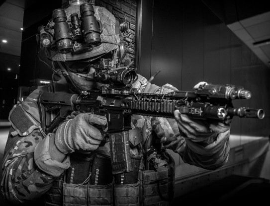 The image is black and white. The image is of a NZSAS soldier holding a weapon, pointed to the right of the image. The solider is in full uniform and wearing glasses, gloves, NVG.