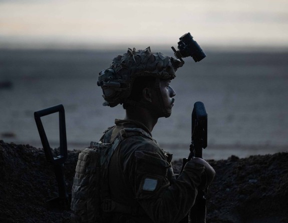 A soldiers keeps watch from a fighting position on a beach looking to the right side of the frame. There is water behind them and the image is almost a silhouette. 