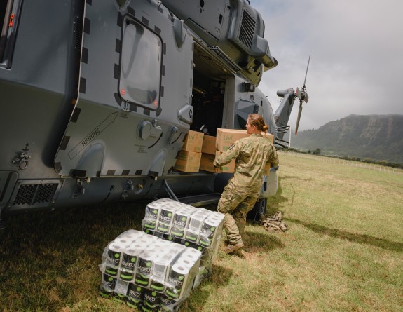 A female military member unloads boxes of toilet paper and water from a grey military helicopter. The helicopter sits on a green field surrounded by large cliffs.