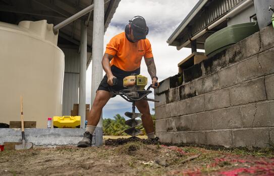 Worker wearing high-visibility clothing and hearing protection operating a motorised earth tool beside a building and water tank.