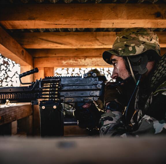 A soldier looks down his weapon while on sentury in a wooden structure with a corrugate metal roof.