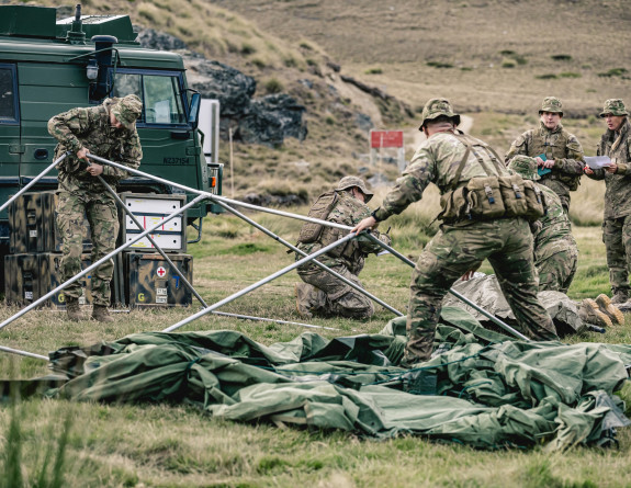 Soldiers in uniform put up a ten. There are other soldiers and a vehicle in the background. 