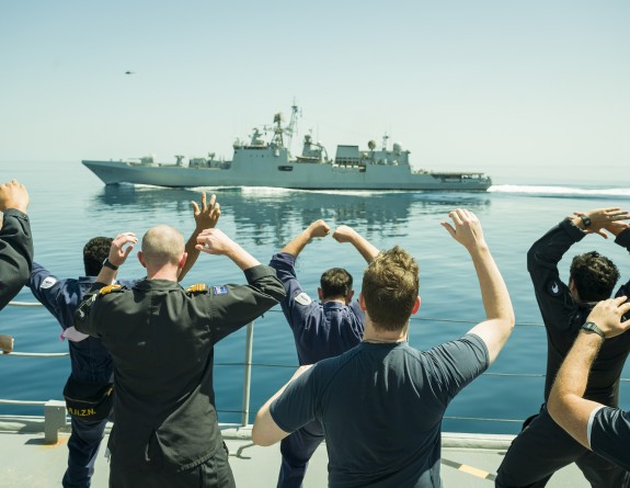 Sailors on a ship at sea, their backs to the camera, hold their hands in the air as they perform a haka to a grey military ship in the background.