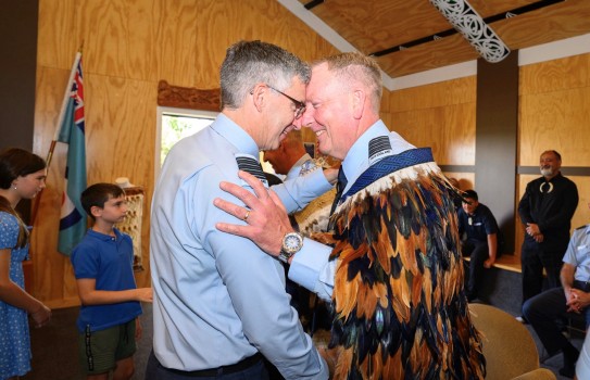Two RNZAF men perform a hongi within the turangawaewae. One wears a korowai, both are smiling while others look on in the background.