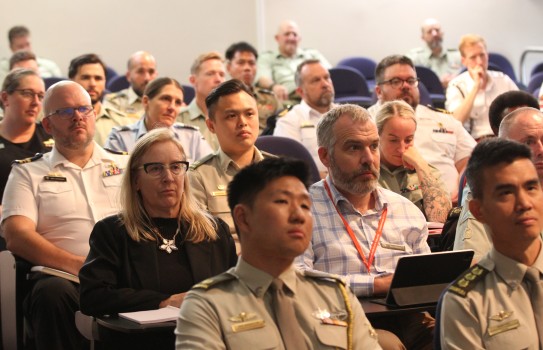 A group of civilian and military, male and female are seated in an auditorium looking toward a single point, presumably a speaker. 