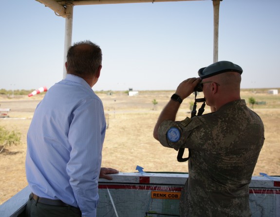Two men stand in a heightened structure shaded by the roof above. One of the men, wearing an army uniform and a green beret, looks through binoculars out into the airfield beyond whilst another man, his back to the camera wears a blue shirt and dress pant