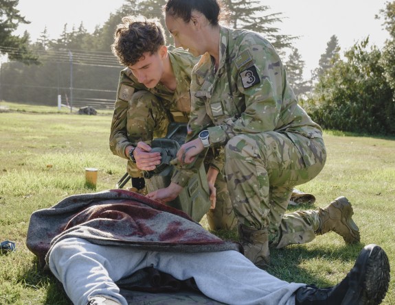 A patient lies on the grass, legs splayed and covered in a blanket that is splashed with simulated blood.  A first aid kit sits in the foreground nearby while a male and female soldiers hold a jerry can together and give water to the patient.