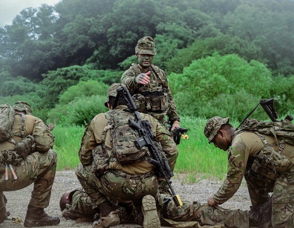 three army soldiers aid another lying down whilst another soldier stands in the front giving instruction. The soldiers wear weapons slung on their backs amongst the grass.