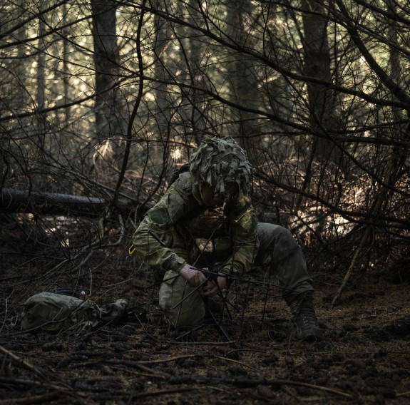 A soldier prepares an aerial underneath the forest canopy, they are camouflaged within the forest.