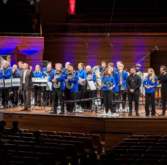 A large brass band dressed in blue jackets and black pants stands on stage with their instruments.