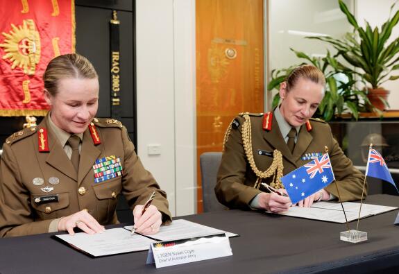 Two Chiefs of Army put pen to paper while sitting at a table with their nations flag front of them. 