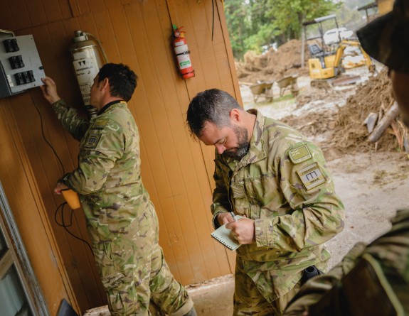 Three military members assess a building's electrics, one looks inside an electrical box, whilst another makes notes on his notepad.  A third person looks on. A digger is positioned to the rear of the building along with piles of tree debris and wheelbarr
