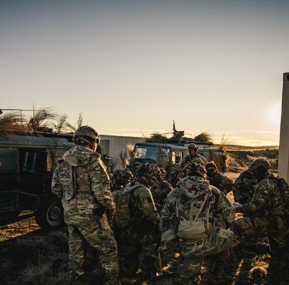 Soldiers congregate at sunrise in front of a couple of small military vehicles.