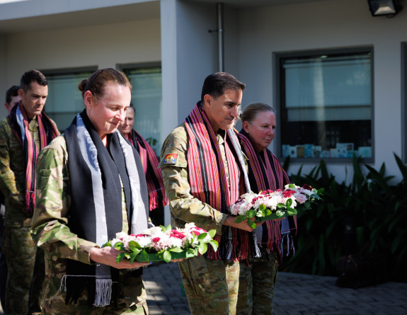 Two uniformed military members hold wreaths and are looking down. There are others around them in uniform. There is a flag to the right of the shot. There is some sunlight on their faces - shining through spaces on a building
