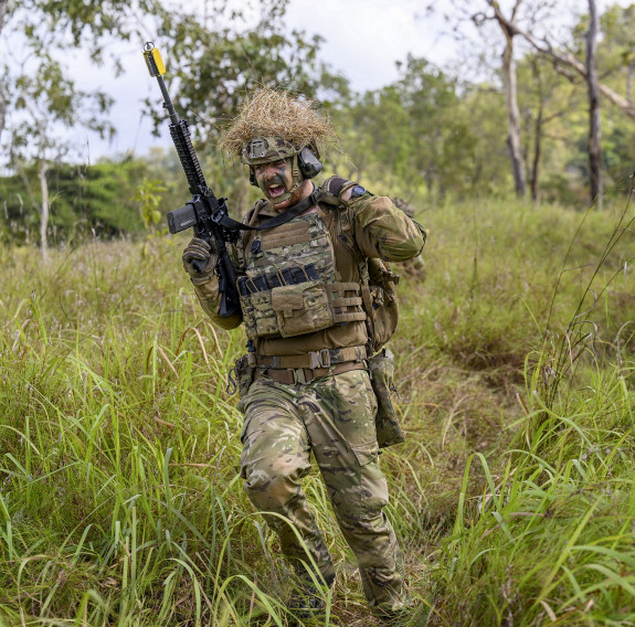 A soldier runs through tall grass towards the left of the frame while holding the MARS-L rifle to the sky. Two other soliders sit low in the grass in the background.