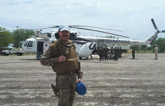 A soldiers stands on tarmac in front of a white helicopter with the word "United Nations" written on the side. He is also wearing a light blue hat.