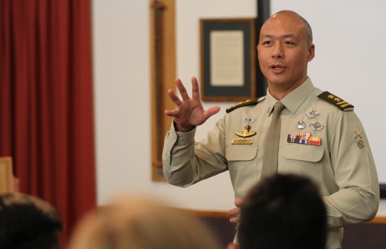 An army officer stands at a lectern speaking to an audience below. Animated and gesturing to the audience as he speaks, the army officer is highly decorated and wears many ribbons and pins on his shirt.
