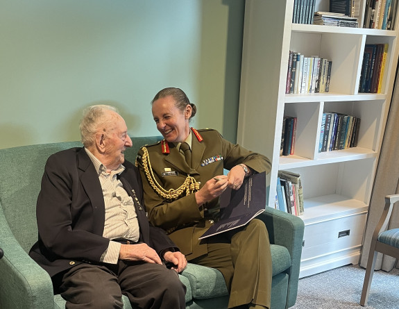 Mr Doug Reid sits with Chief of Army Major General Rose King on a two-person sofa next to a bookshelf, as they both laugh.
