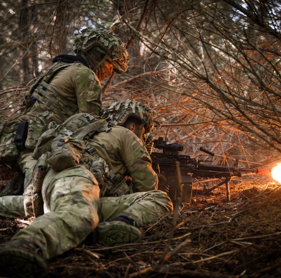 Two soldiers side by side, one prone and the other seated whilst firing a weapon 