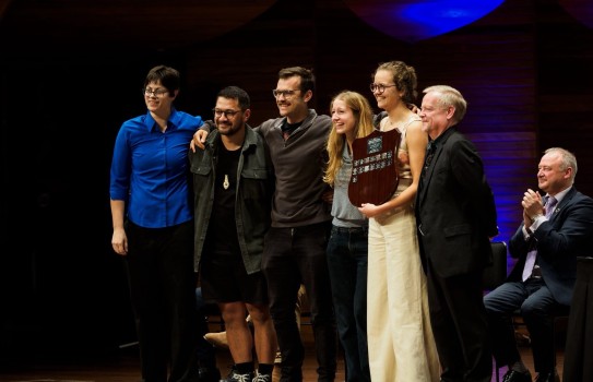 A small group of people in various dress ranging from casual to formal stand on stage, arms around one another smiling and holding a shield.