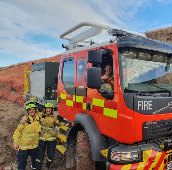 Two people with smiles and arms around each other stand in front of a NZDF firetruck, with another person in the drivers seat.