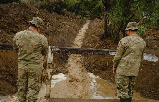 Two soldiers stand on a small bridge looking down a waterway that runs between two freshly scraped dirt.