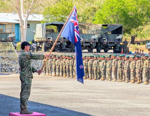 A New Zealand soldier stands in uniform, holding the NZ flag in front of various military personnel, standing in a line. They are all in the distance and slightly blurred out of focus. 