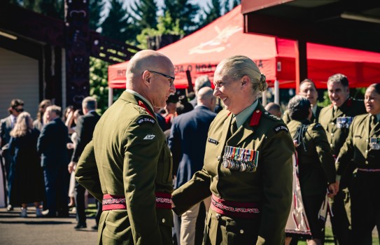 Two Army officers, a male and female greet one another whilst wearing ceremonial uniforms. The female wears medals on her left chest and smiles at her male counterpart. People in uniform gather behind them near a red marquee.