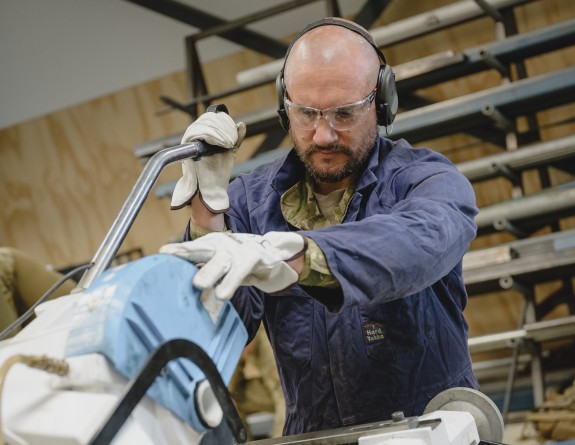 A male, middle aged with moustache and a beard wears PPE of safety glasses, hearing protection and safety gloves while he uses a industrial drop saw.  He wears blue overalls over his green camouflage uniform.