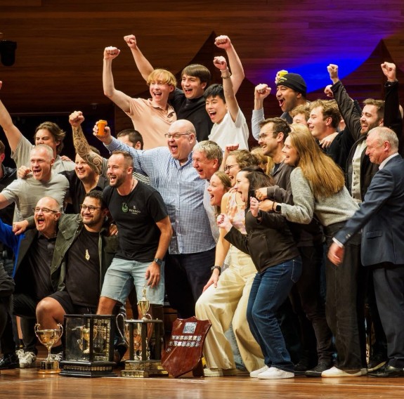 A group of people stand around a number of trophies, arms in the air, smiles, jubilantly celebrating their wins.