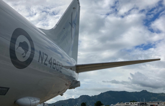 The tail of the RNZAF P8-A Poseidon with a grey livery, the kiwi roundel and the code 'NZ4802'. The aircraft sits on the tarmac in Fiji under scattered clouds.