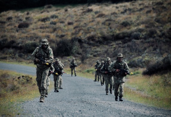 A group of Army Soldiers patrol dressed in camouflaged uniforms and carrying weapons down a gravel road with the tussock hills behind.