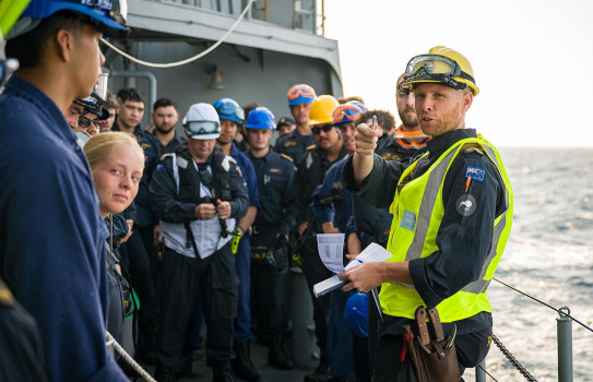 Some Navy personnel wear coloured vests that indicate their role, other wear regular Navy uniform while most wear hard-hats and have protective goggles on their helmets as they talk through their plan.