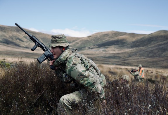 A soldier holds a weapon as they move through tussocks in front of rolling hills.