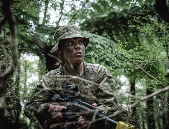 Private Zara Reid takes part in regular training with Wellington Company, 5/7 Battalion, RNZIR. She stands in a forrest, alert of her surroundings donned in full camouflage.