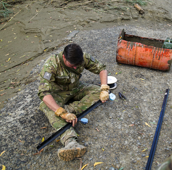 A soldier sits on the ground next to a river, one leg straight, and the other tucked underneath it as he works to build a pulley system using a metal beam, and an oil drum.