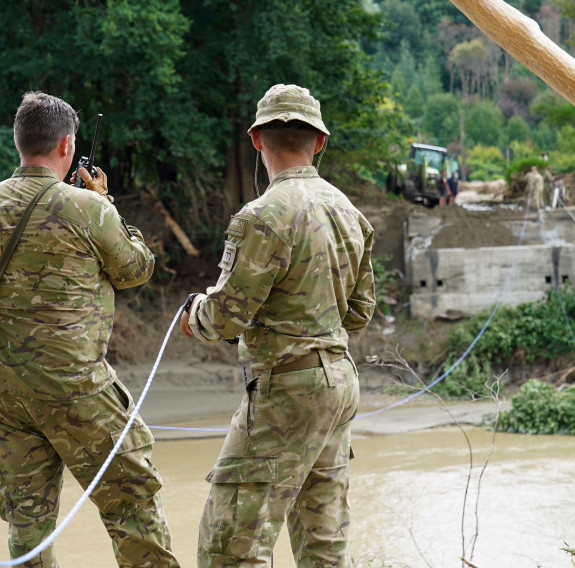 Three soldiers stand on the bank of a river where a bridge once stood. On the other side of the river, a small part of the bridge remains but the rest is gone. Debris from the high flood waters is caught in trees and on the sides of the river.