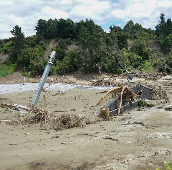 A house is engulfed is mud, sand and debris from the storm, only the edges stick out from the sludge. There is a ridge in the background with trees on it, there are slips where the land has given away which span the entire image.