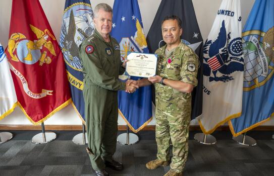 Two men shake hands while handing over a plaque award. They are standing in front a of multiple flags.