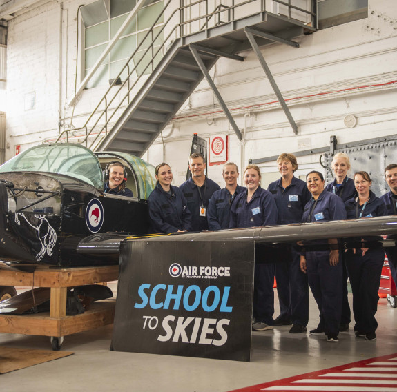Teachers stand behind the wing of a small aircraft in a hanger.