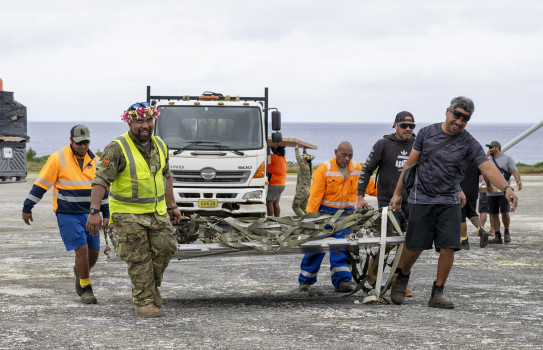 Four people and a RNZAF aviator lift a large metal object that has material netting on top.