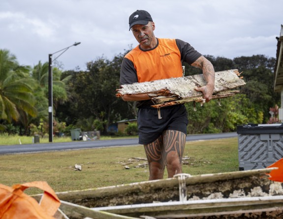 A soldier in a high visibility tshirt and shorts walks towards the camera carrying some rugged pieces of wood.