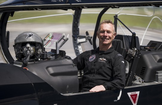 A male wearing a black flying suit sits inside the cockpit of a small aircraft with the hood open. His hand rests on the side of the aircraft whilst looking at the camera.