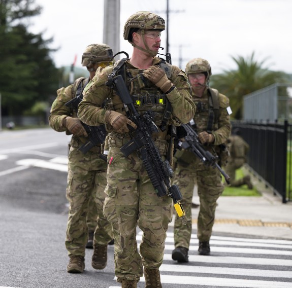Three soldiers cross a zebra crossing in full Army uniform while holding rifle systems
