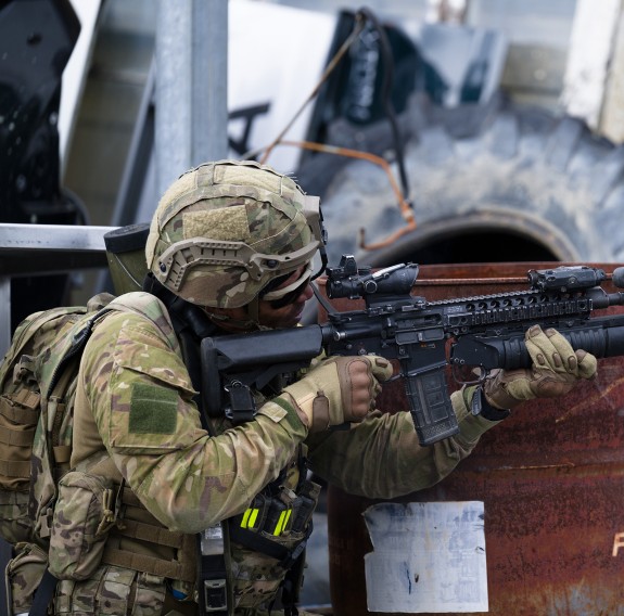A soldier looks through the sights of a MARS-L as they lean against a barrel with a large tyre and panels of wood are stacked in the background.