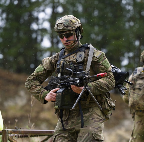 A soldier holds his weapon as he walks from right to left. In the background other soldiers are talking in front of large trees.