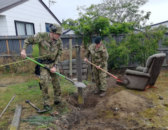 Two New Zealand Army soldiers dig holes in the dirt as part of fence building. It's a grey day and in the background, you can see a house over another fence. 