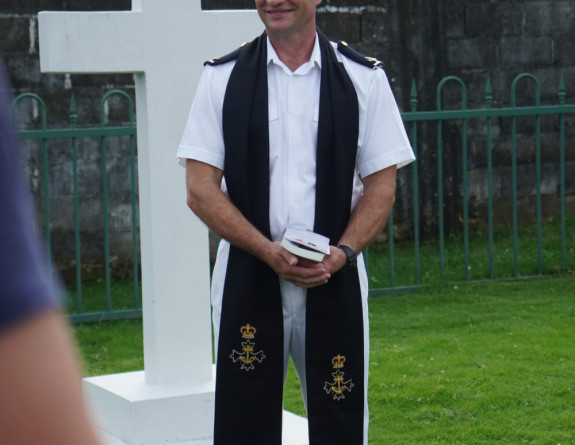 Chaplain Lloyd Salmon smiles as they wear dress whites, standing in front of a white cross.